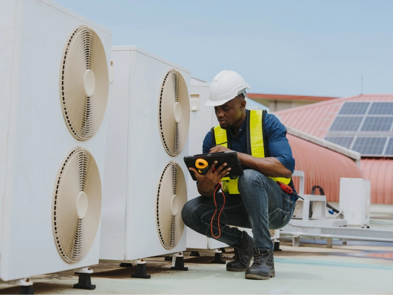 Man in a hard hat and safety vest kneeling down next to a heat pump