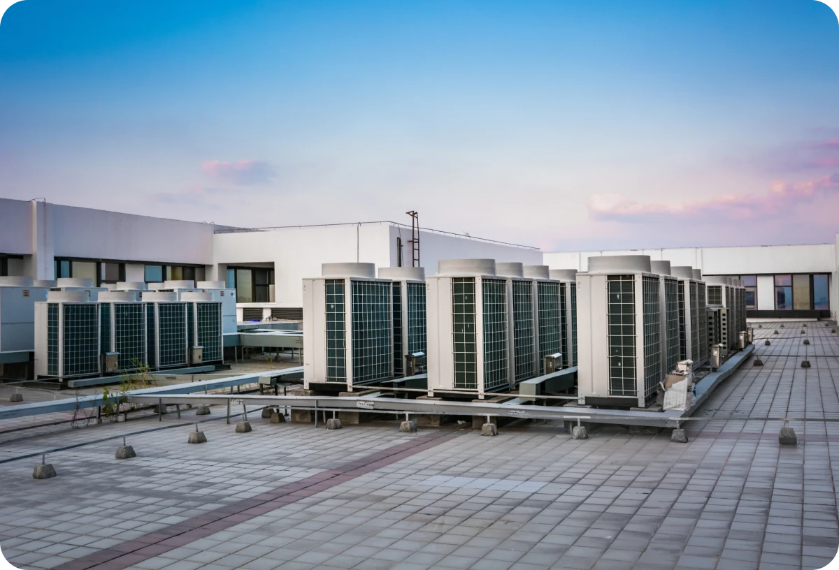 Large building with a lot of air conditioning units on its roof, surrounded by plants and poles