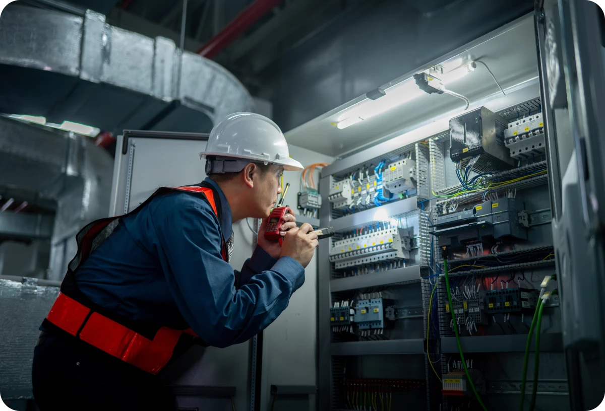 Man in a hard hat and safety vest working on an electrical panel, holding a device in his hand