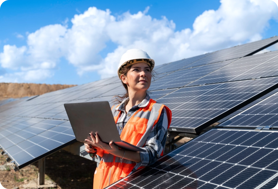 Woman in an orange vest and hard hat standing in front of solar panels, holding a laptop in her hands